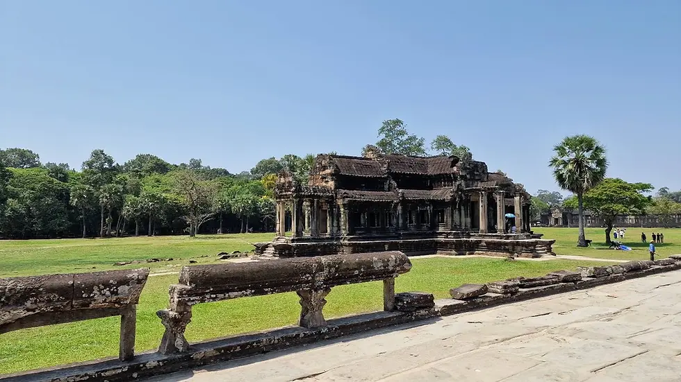 Ancient temple with stone carvings set in a lush green field, under a clear blue sky. People with umbrellas nearby, trees in the background.