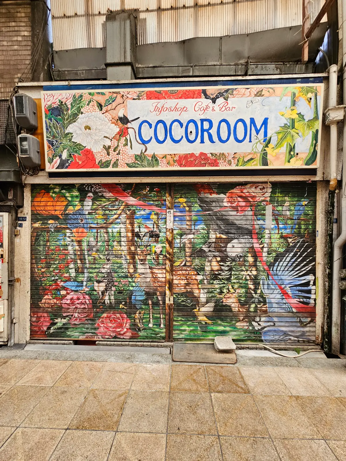 A shopfront with a sign reading "COCOROOM" above a colorful mural featuring flowers, birds, and lush plants. The artwork covers the entire closed shutter, giving the area a lively, vibrant appearance.