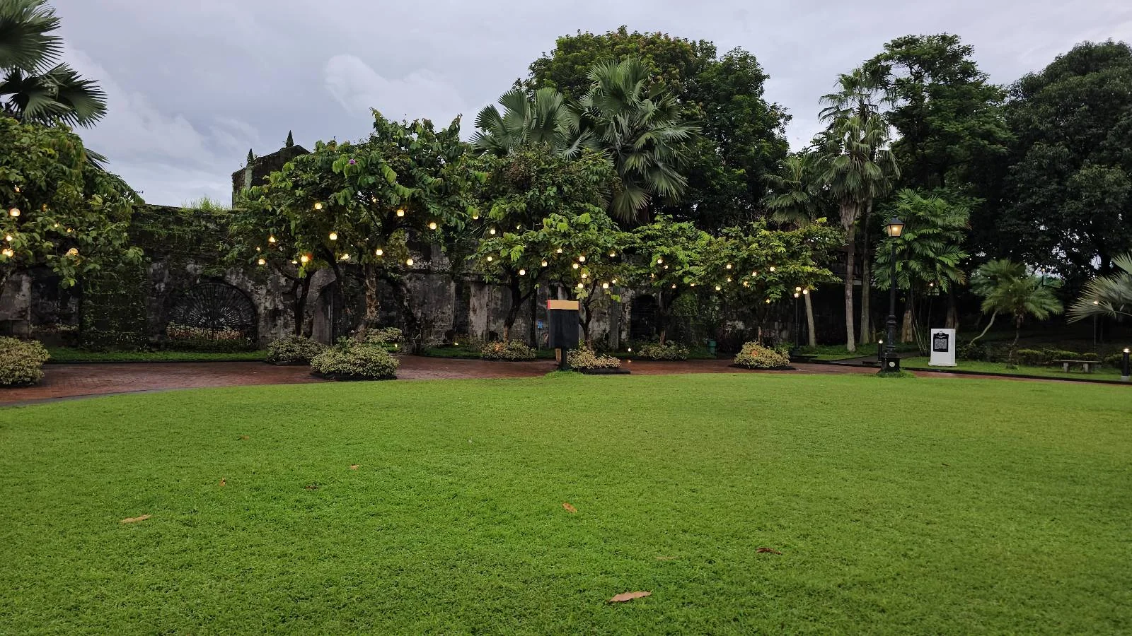 A lush green garden with manicured grass is surrounded by tall trees adorned with string lights. Overcast sky adds a soft, diffused light to the scene. A lamppost stands in the center, and a pathway is visible in the background.