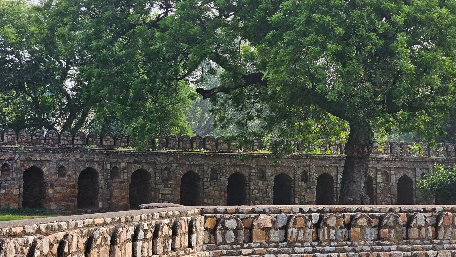A historic stone wall with a series of arches stands under a large, leafy tree. The wall is made of weathered stone blocks, blending naturally with the lush greenery surrounding it. A pathway of similar stone is in the foreground.