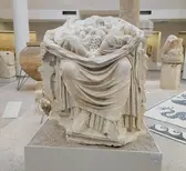 Ancient Roman sculpture of figures enveloped in draped robes, displayed in a museum setting with other artifacts.