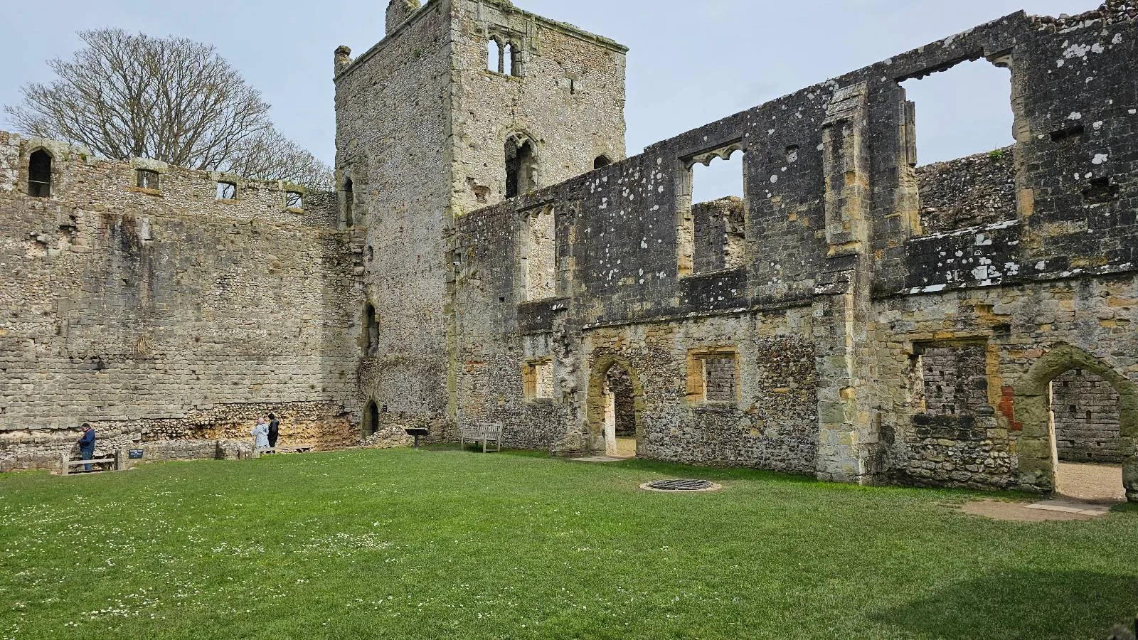Ruins of an old stone building with tall, broken walls and empty window frames surrounding a grassy courtyard; a few people are visible near the base of the structure.