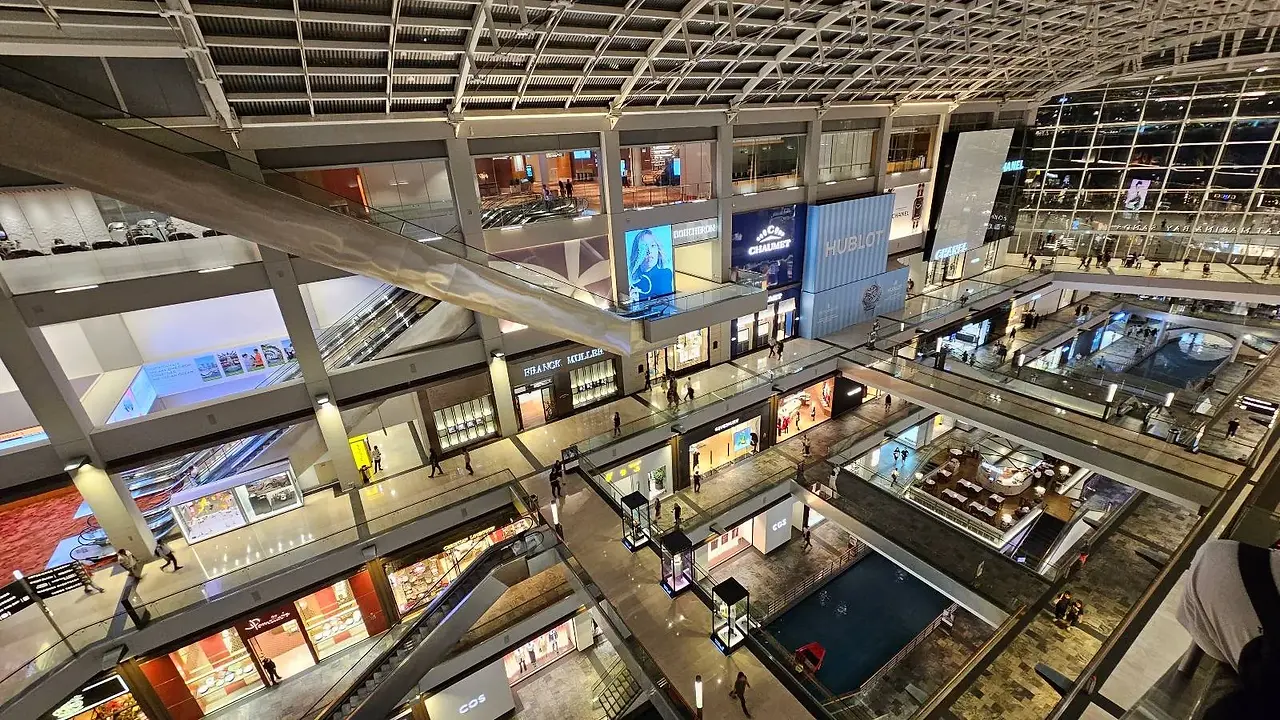 A spacious, multi-level shopping mall with brightly lit store fronts and escalators. The modern architecture features a glass ceiling and large digital screens displaying advertisements. Shoppers are visible on various levels.