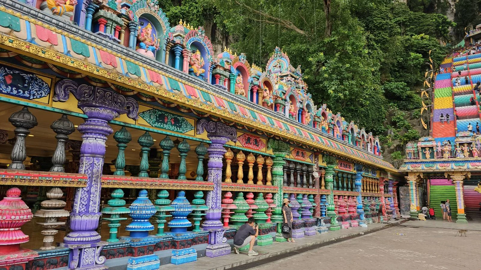 Colorful temple façade with intricate sculptures, vibrant columns, and rainbow stairs. People interact and admire the details. Greenery surrounds.