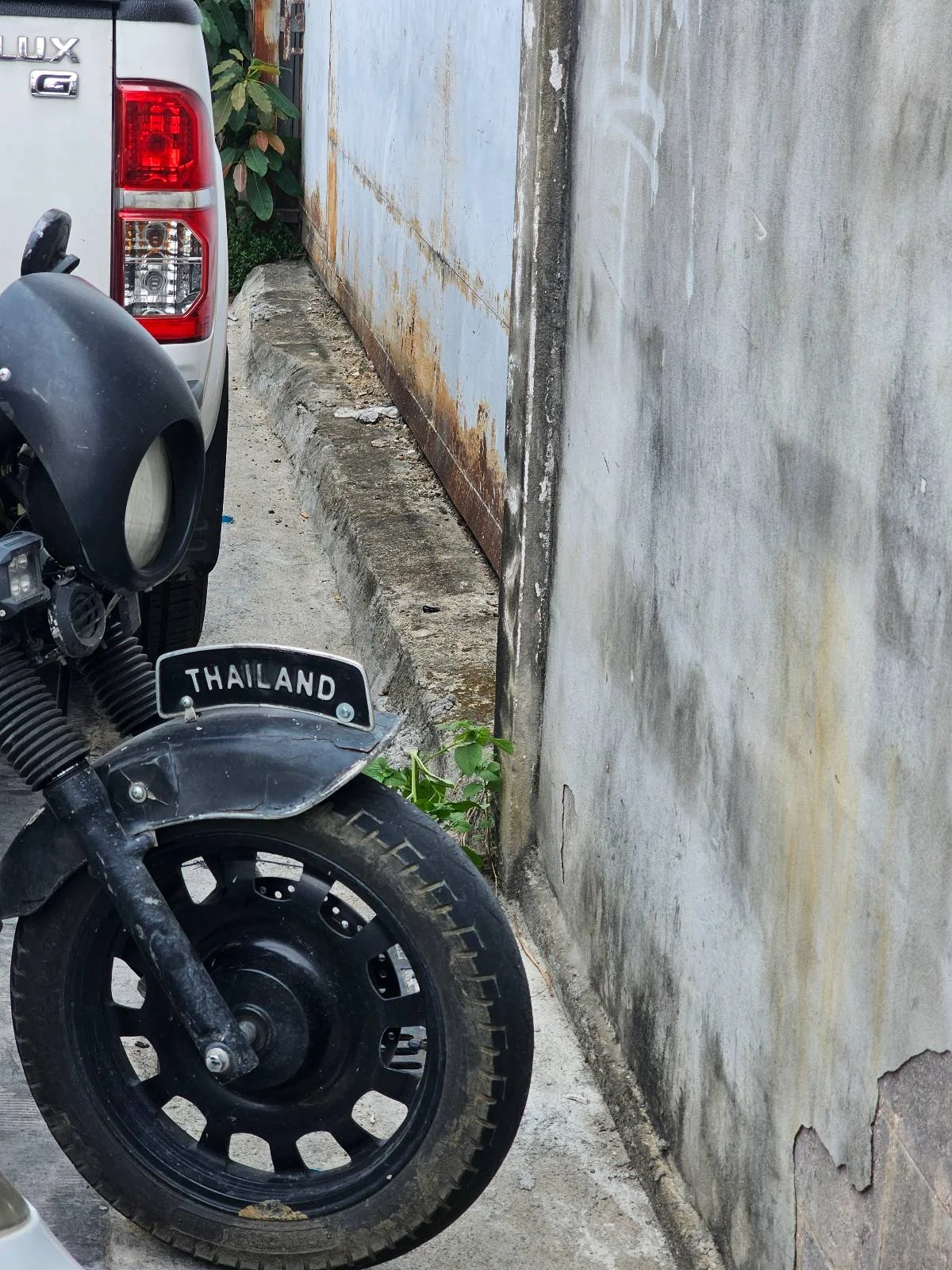 A black motorcycle with a "Thailand" license plate is parked next to a white vehicle. The background features a faded, weathered wall and a narrow concrete path.