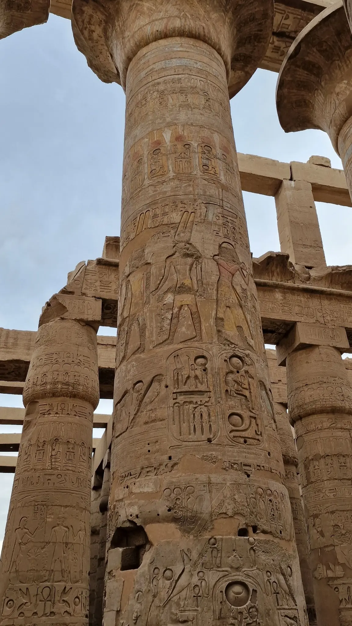 A tall, ancient stone column with intricate carvings and hieroglyphics stands among other ruins, under a partly cloudy sky.