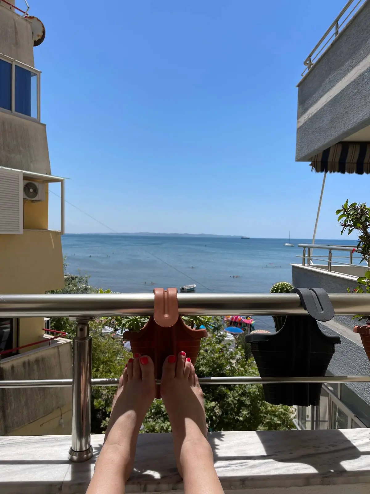 Person relaxing on a balcony with feet up on a railing, overlooking a calm sea under a clear blue sky, with buildings and greenery on either side.