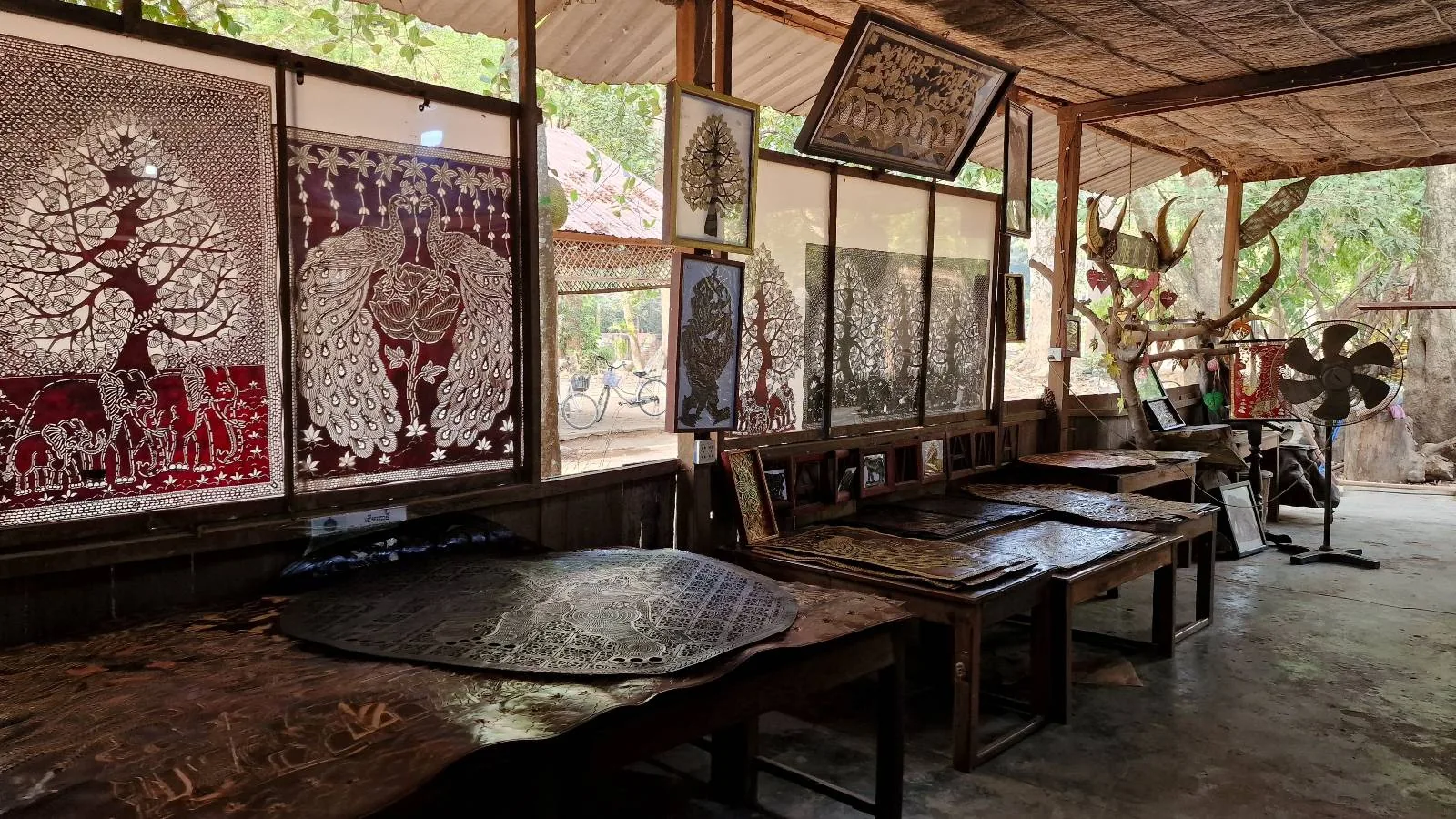 A rustic workshop interior displays intricate metalwork art pieces. Several tables hold various metal designs, including one shaped like a tree, under a wooden roof with natural light filtering through.