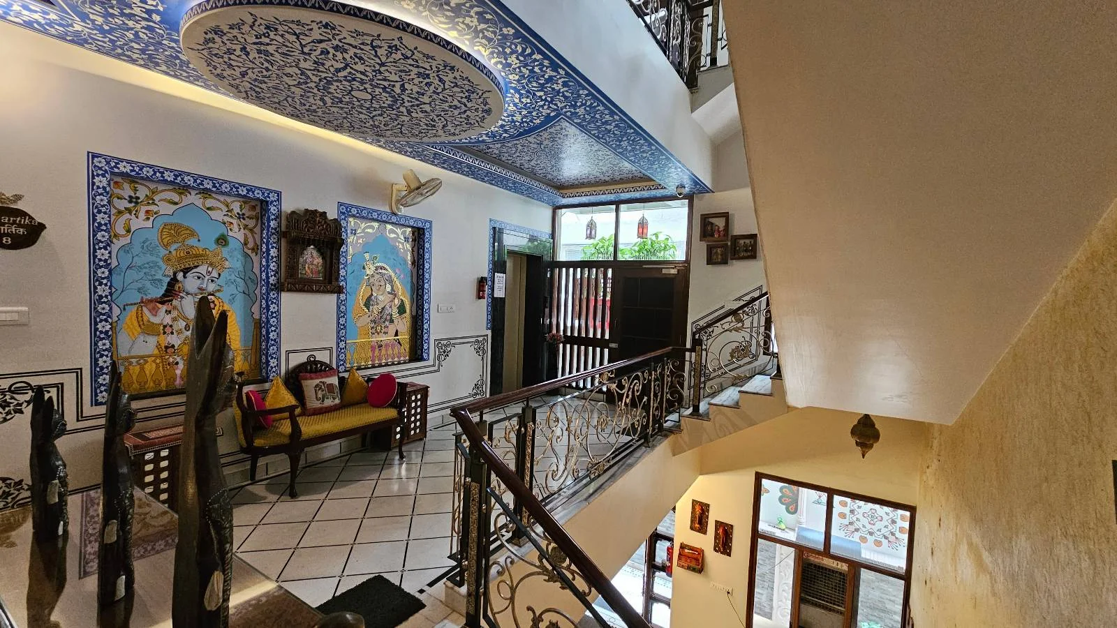A vibrant interior of a house with ornate blue ceiling art, colorful wall paintings, decorative railings, and a cozy seating area along the wall. Natural light enters through windows, creating a warm and inviting atmosphere.