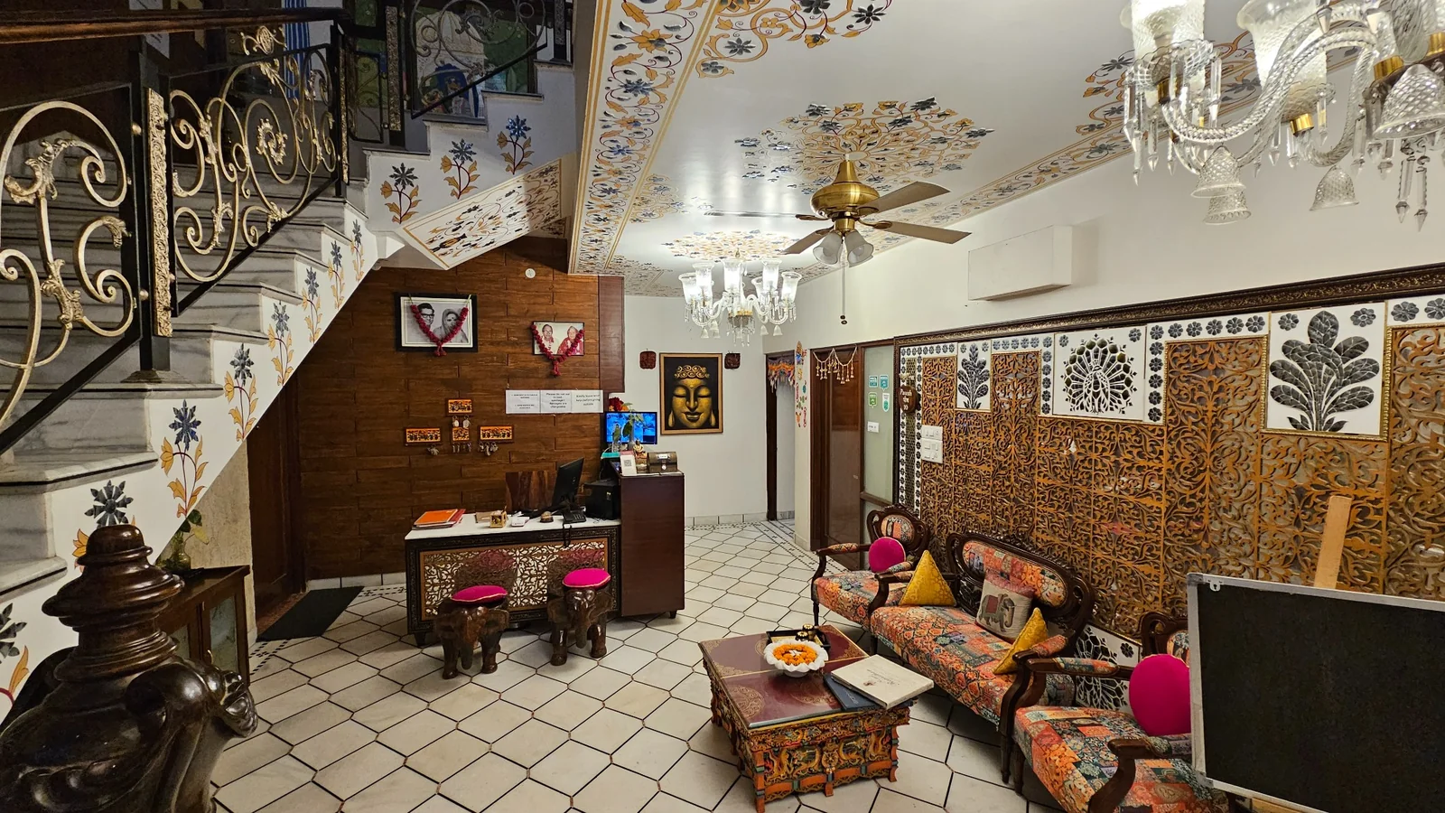 A cozy, ornately decorated living room with patterned furniture, a carved wooden coffee table, wall art, chandeliers, and a staircase with intricate railings. The ceiling has colorful designs and a ceiling fan.