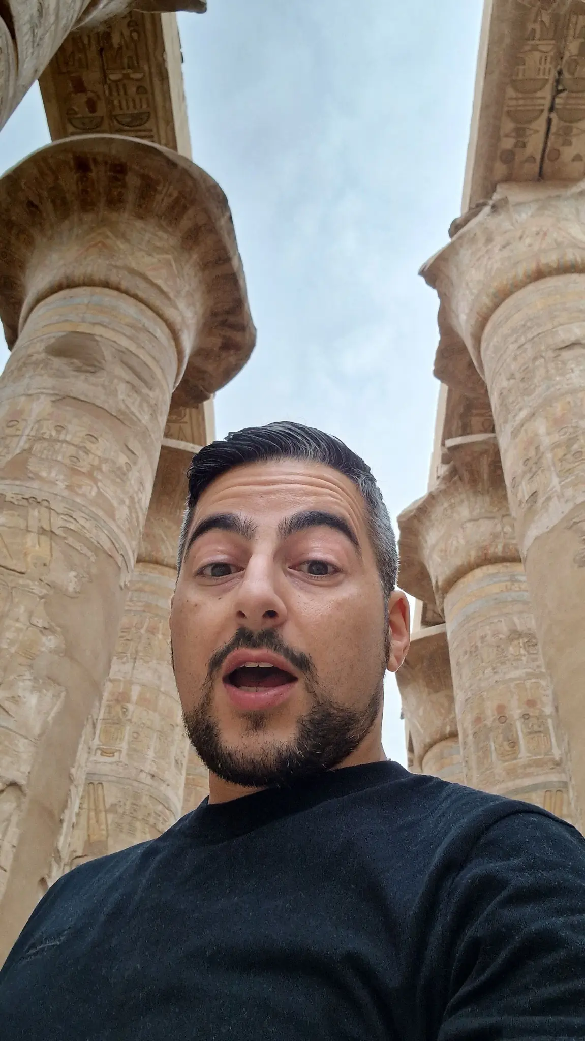 A person with a beard and short hair stands in front of large, ancient stone columns, wearing a black shirt, looking slightly surprised. The sky is cloudy.