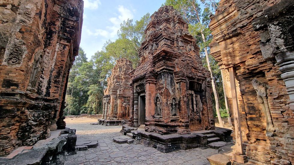 Ancient brick temple ruins surrounded by lush trees under a blue sky. Intricate carvings adorn the weathered structures.