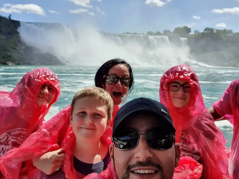 Group wearing red ponchos smiling in front of a waterfall. Clear sky, water splashing in background. Energetic and joyful mood.