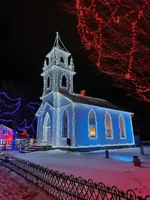 A church illuminated with white and blue Christmas lights stands against the nighttime sky at Ontario's winter festival. Nearby trees are adorned with red and blue lights, while a blanket of snow adds to the festive atmosphere in Upper Canada Village.