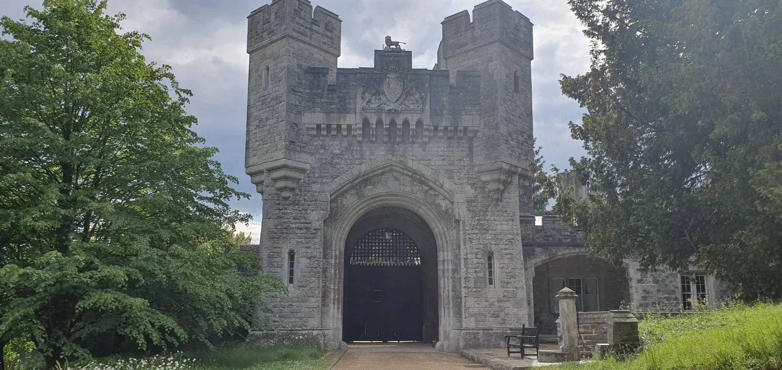 A stone castle entrance with two cylindrical towers flanking a large archway under a cloudy sky. Lush greenery surrounds the structure, with trees on both sides. The path leads through the open archway.