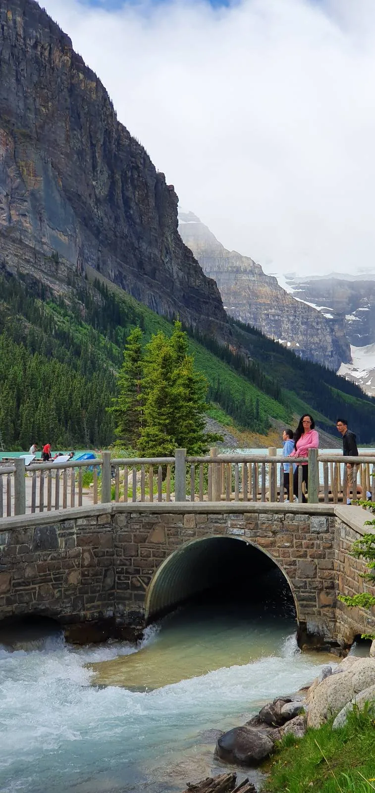 A stone bridge stretches over a flowing river with a couple standing on it, surrounded by lush green trees and towering mountains partially shrouded in clouds.