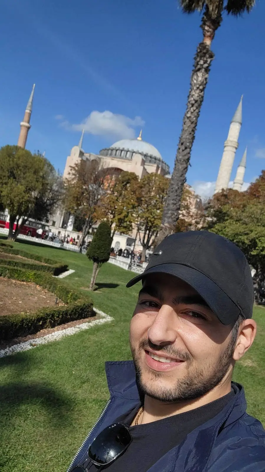 A person in a black cap smiles in the foreground with a grassy area, trees, and a historic building with minarets in the background under a clear blue sky.