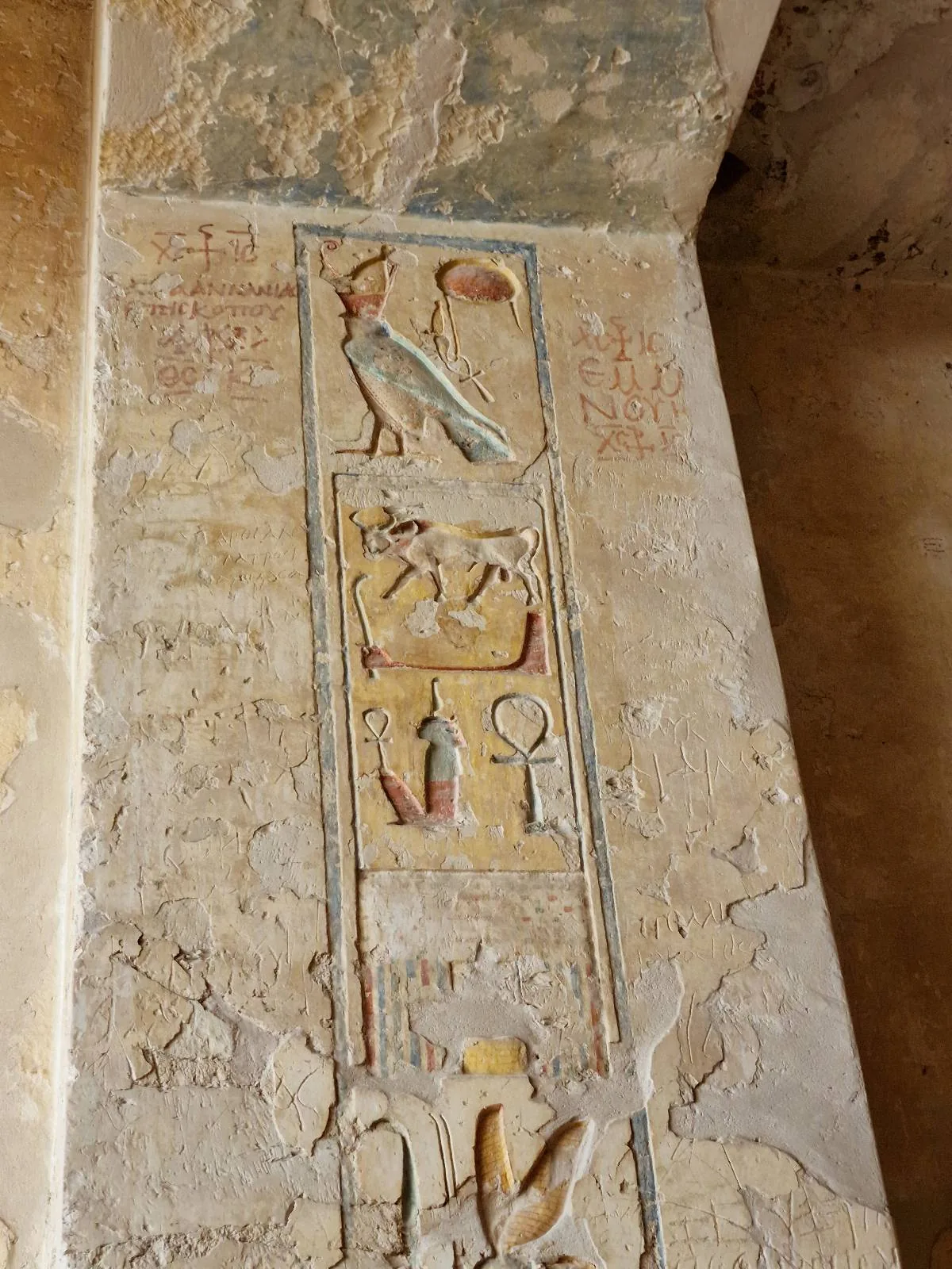 Ancient Egyptian hieroglyphics on a weathered stone wall, including symbols like a bird, an ankh, and other traditional figures in vertical alignment.