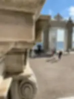 Stone archway overlooking the sea, framed by ornate architectural details. People walk nearby under a clear blue sky.