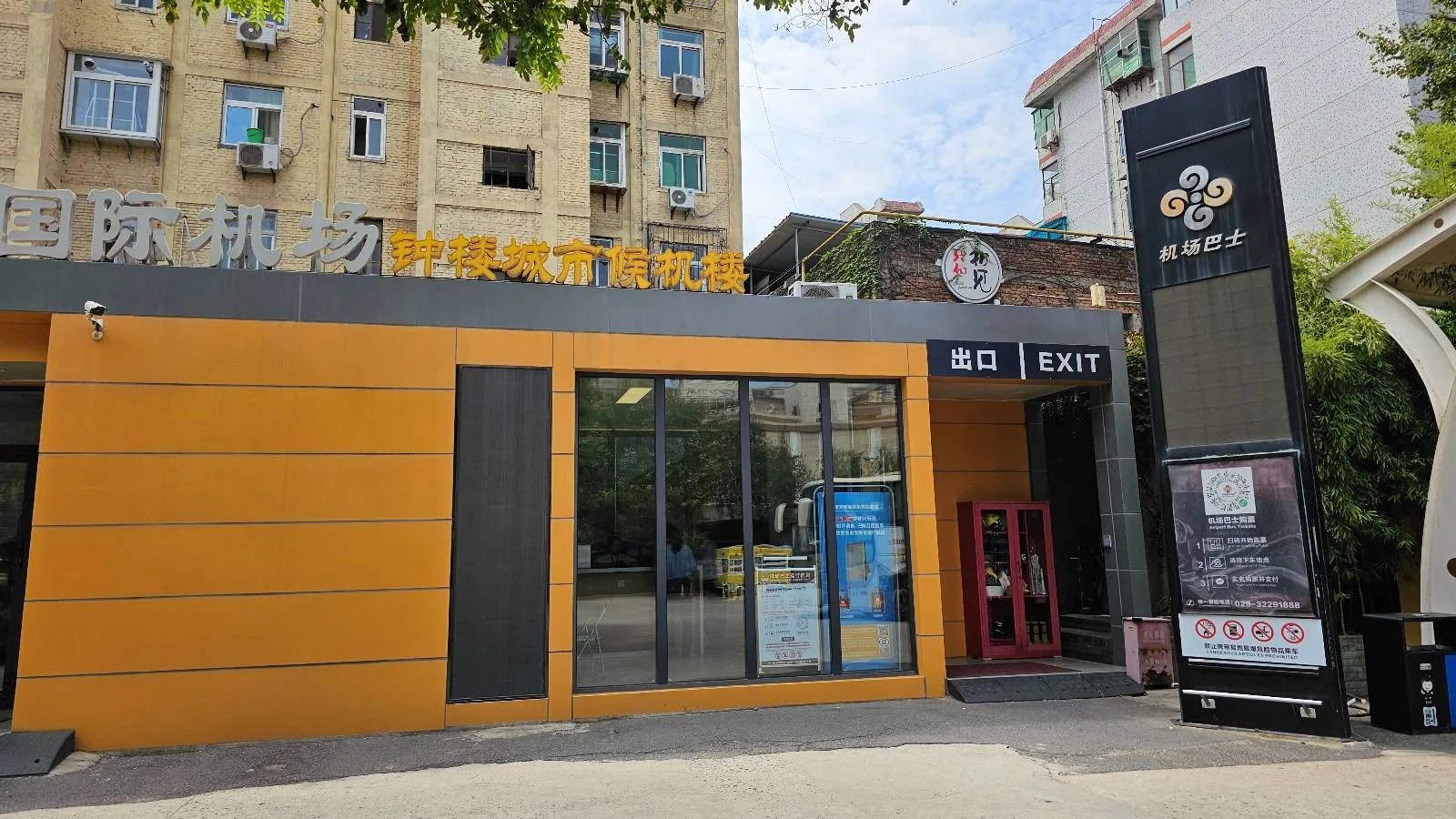A building with a yellow and black exterior features large glass windows, a red door, and an exit sign. There’s a tall black sign with white and orange text near the entrance, and apartment buildings in the background.