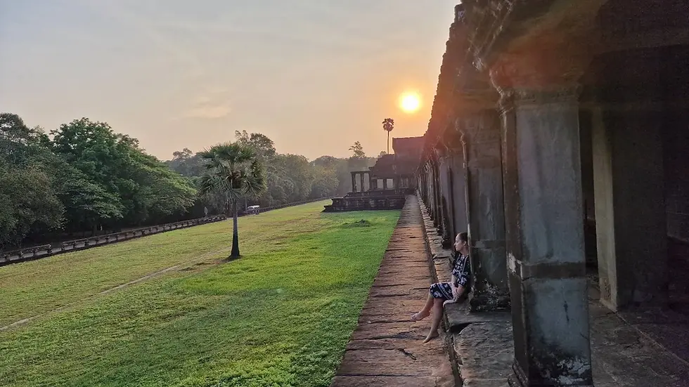Person sitting on ancient stone walkway at sunrise, next to green lawn and trees. Sun creates serene ambiance. Rust-colored pillars line the path.
