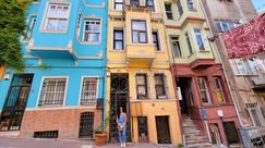 A person in blue jeans and a white shirt stands before a bright yellow building in Istanbul, surrounded by colorful buildings in blue, green, and red on a vibrant city street—an unforgettable place in the city's cultural capital.