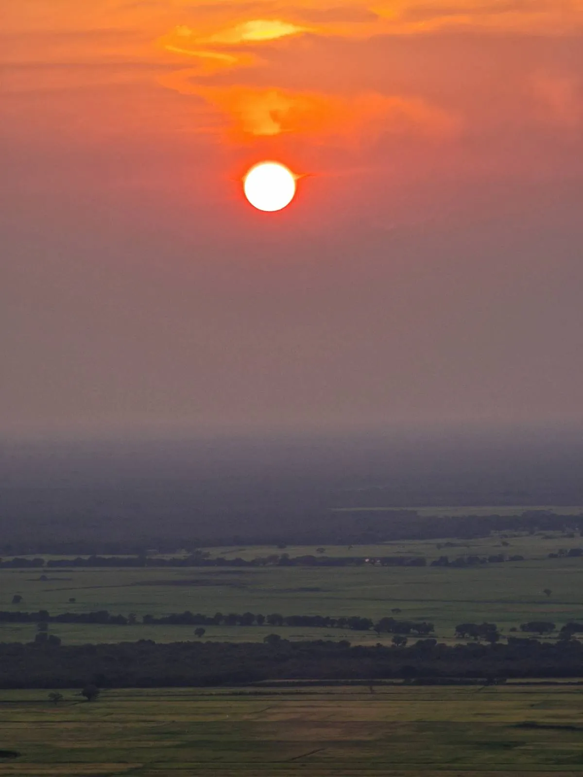 The image shows a vibrant sunset with an orange sun low in the sky, casting a warm glow over a misty landscape of green fields and distant trees.