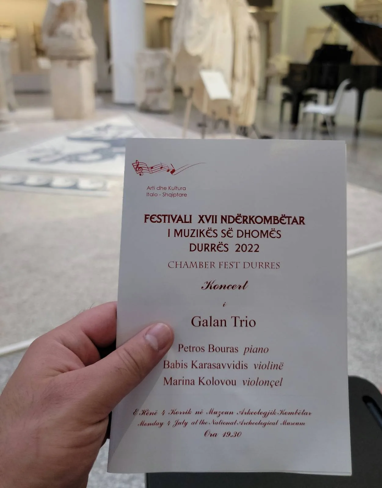 A person holds a concert program in a museum setting with classical sculptures visible in the background. The program mentions a gala trio and lists several composers: Brahms, Gliere, Popper, and Shostakovich. A piano is also visible in the scene.