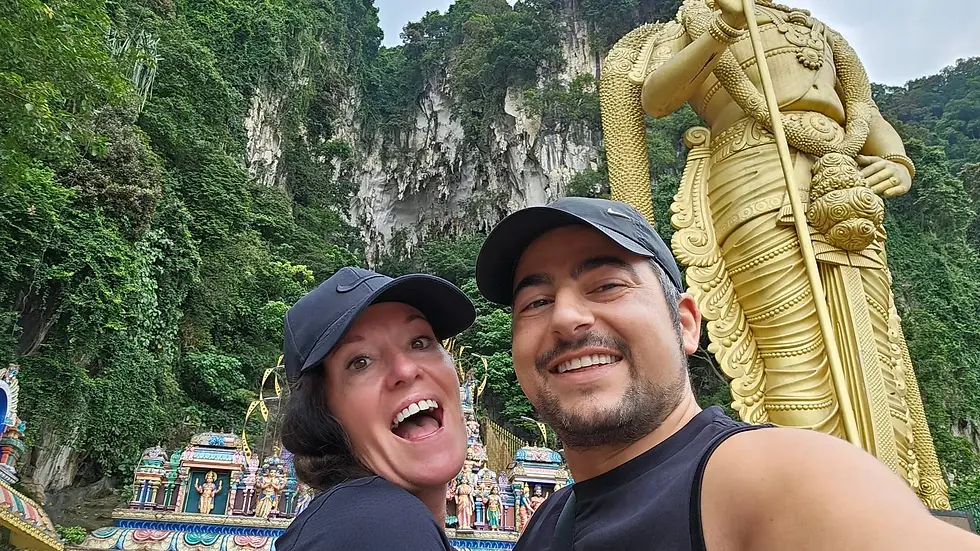 Smiling couple in black caps take a selfie near a large golden statue, with lush green cliffs and colorful temple structures in the background.