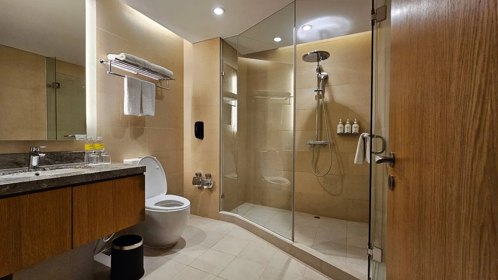 Modern bathroom with a glass-enclosed shower, wall-mounted showerhead, toilet, sink with a large mirror, wooden cabinets, and beige tiles. Towels are neatly hung on the wall, and toiletries are visible in the shower.
