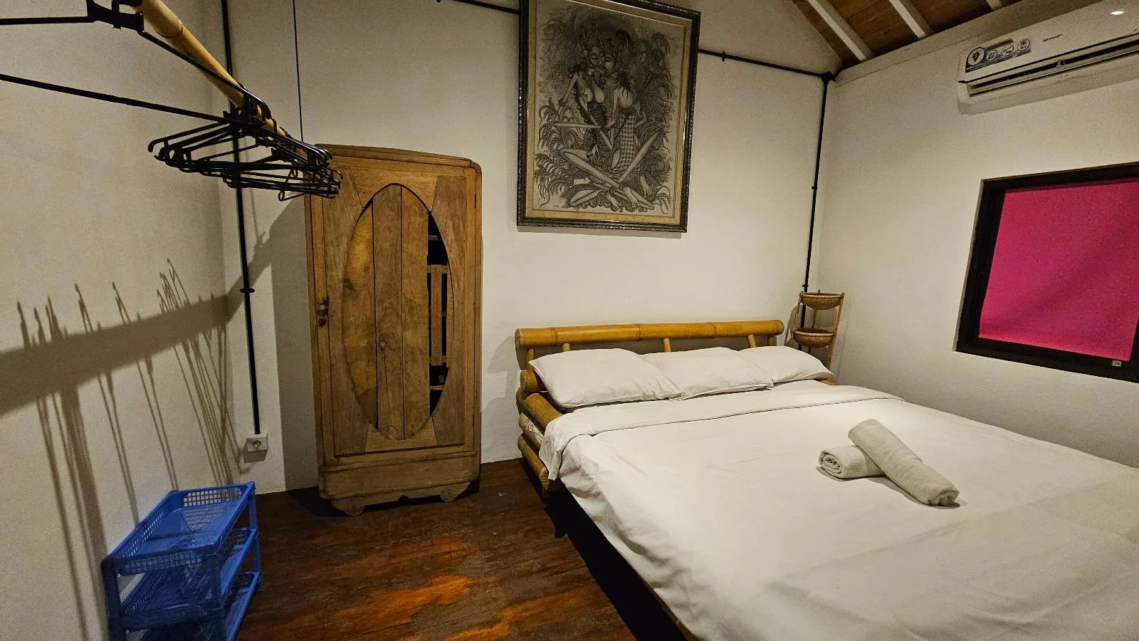 A cozy bedroom with a wooden bed topped with white bedding and a neatly folded towel. A wooden wardrobe stands in the corner next to a hanging rack. The walls are adorned with a framed picture and there's a window with a pink curtain.