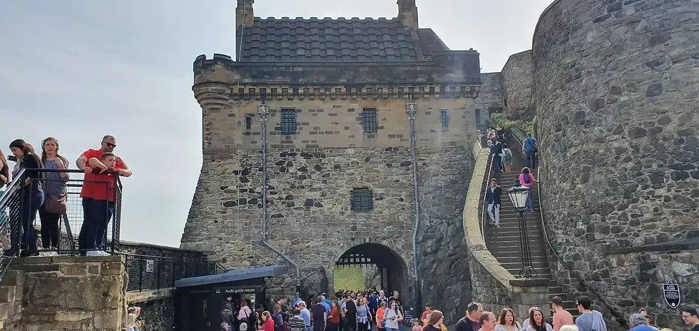 Crowds explore a historic stone castle with stairs and archway. People gather near "Audio guide return" sign. Sunny day, lively mood.