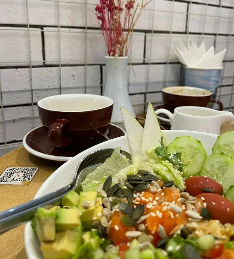 Plate of avocado, cucumber, tomato and seeds accompanied with a brown cup of coffee and a vase of red stem flowers.