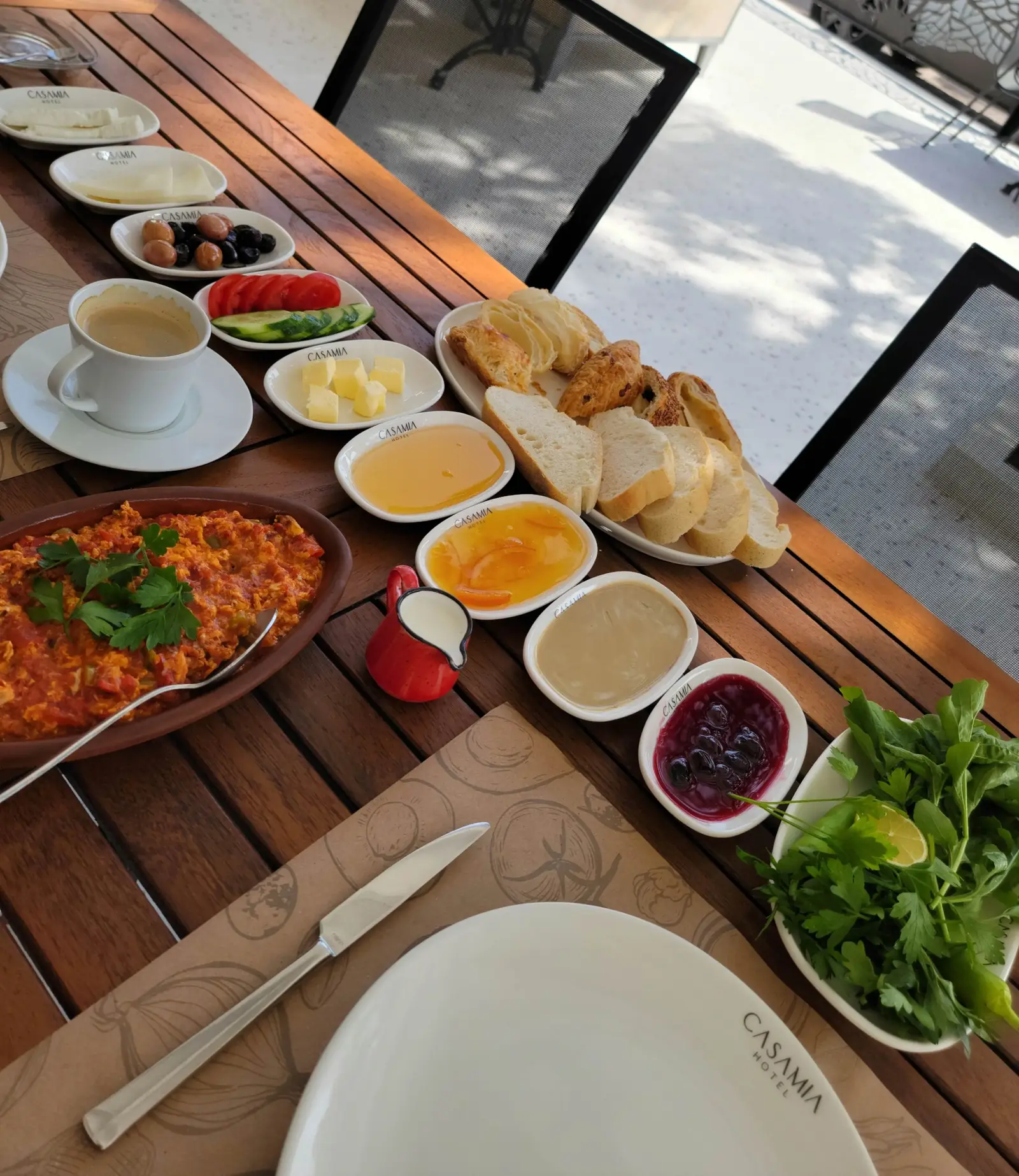 A table with various breakfast items, including sliced bread, spreads, greens, cheese, a coffee cup, and a pan of cooked eggs, set outdoors.