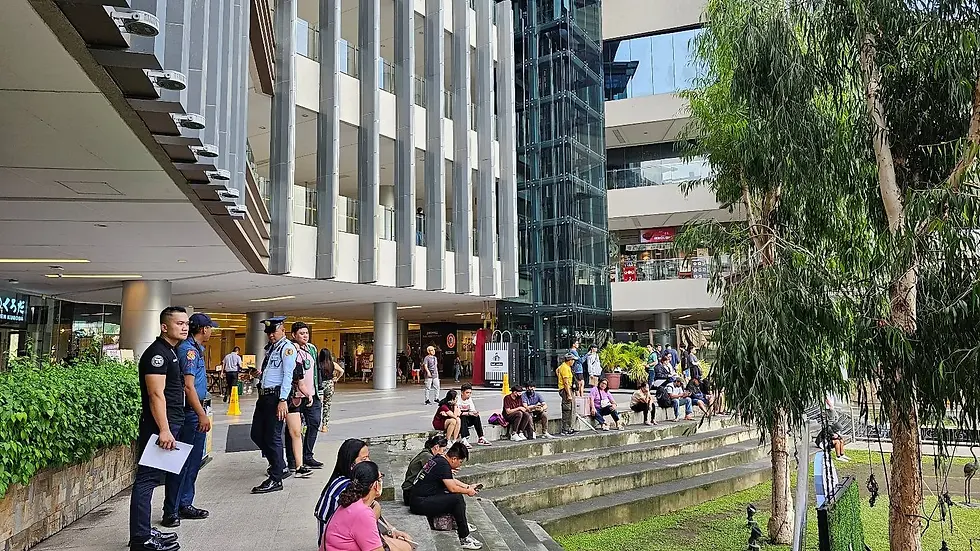 People sit and stand near a modern building with large windows. Security officers watch. Trees and greenery in the foreground.
