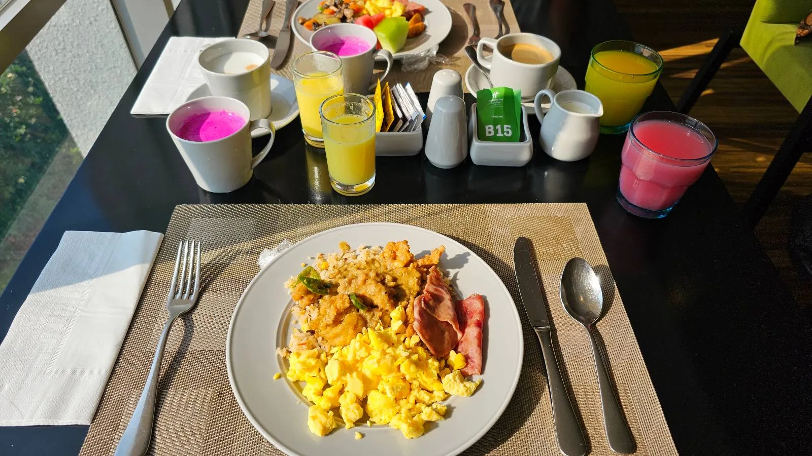 A table set for breakfast with two plates. The front plate has scrambled eggs, bacon, and hash browns. Drinks include coffee and colorful juices. There are condiments and a fruit bowl. Sunlight is streaming in through a window.