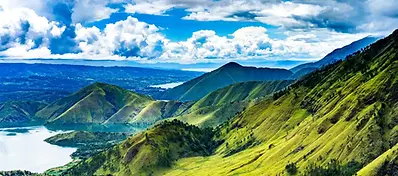 A sweeping view of rolling green hills and mountains surrounding the deep blue waters of Lake Toba under a blue sky with fluffy white clouds.