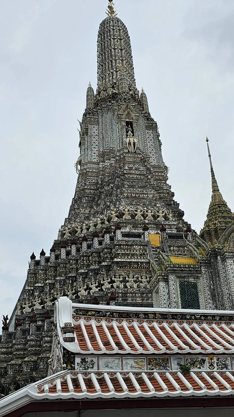A detailed view of Wat Arun, a historic Buddhist temple in Bangkok, Thailand. The temple's central spire is intricately decorated with colorful porcelain tiles and stands against a cloudy sky. A tiled roof is visible in the foreground.