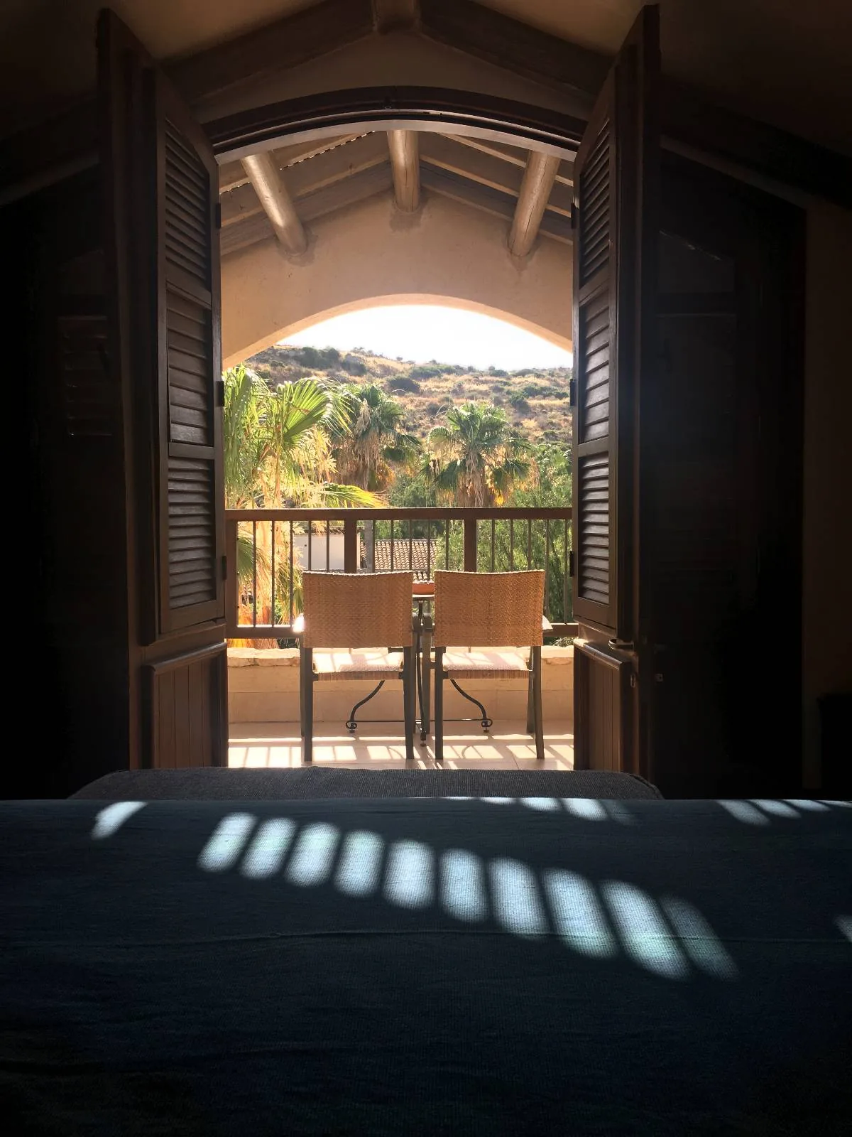 A cozy room with sunlight streaming through wooden shutters. A small balcony is visible, featuring two chairs and a table. Beyond the balcony, a scenic view of hills and trees is partially visible. The atmosphere is serene and inviting.