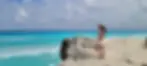 Woman in sunhat and pink dress stands on a rocky beach beside turquoise sea under a cloudy sky.