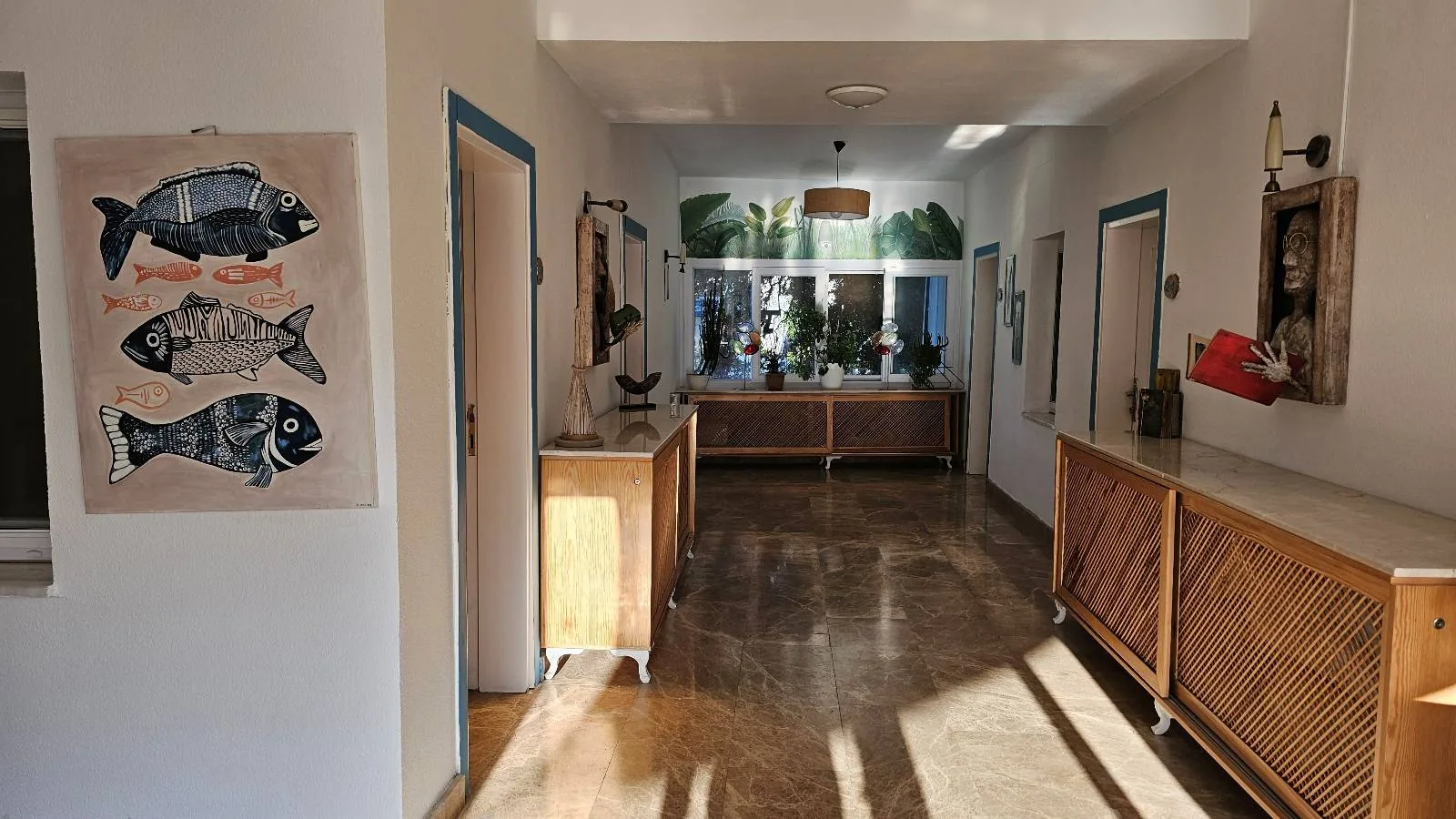 Hallway with fish artwork, wooden cabinets, and a sculpture holding a red book. Plants by sunlit window, walls and floor in beige tones.