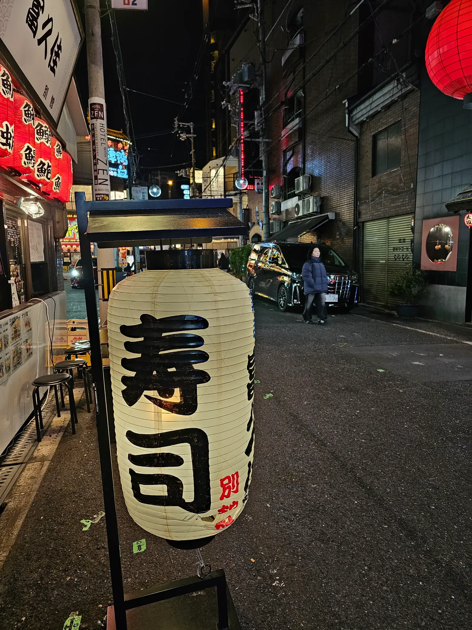 A Japanese street at night with a large lantern in the foreground displaying the kanji for "sushi." The street is lined with buildings, neon signs, and a few people and bicycles in the background.
