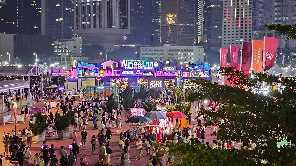 A bustling outdoor night market with colorful vendor stalls and bright signs, including one for "Wine & Dine." Many people are walking around, with tall city buildings illuminated in the background.