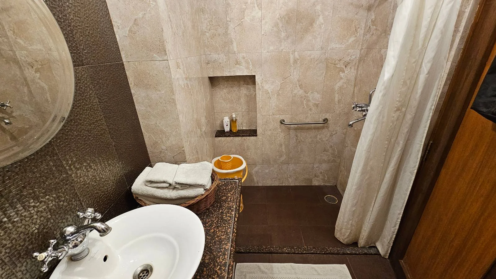 A small bathroom with beige tiled walls, a white sink, a towel basket, a shower area with a grab bar, a soap niche, and a white shower curtain.