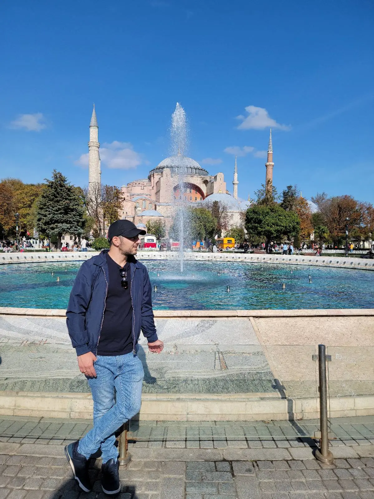 Person in casual attire standing by a fountain with a historic building and minarets in the background under a clear blue sky.