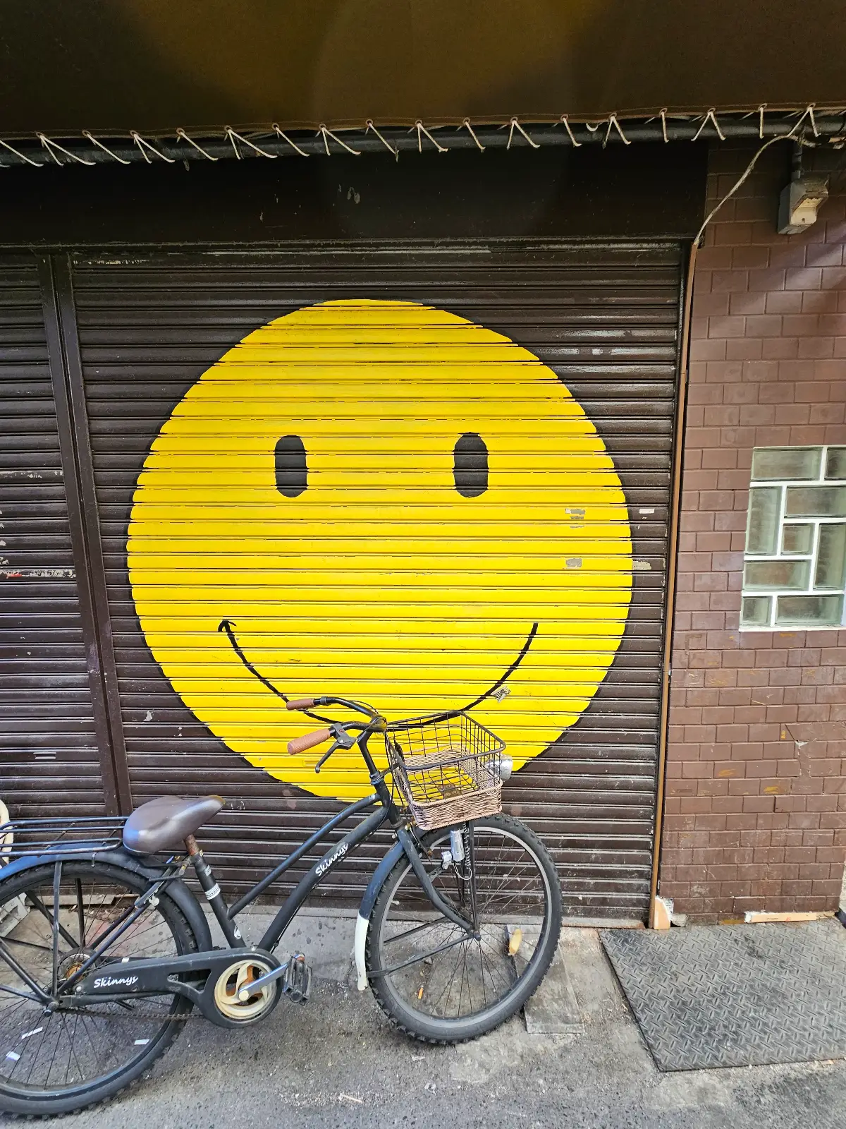 A black bicycle with a front basket is parked in front of a metal shutter painted with a large yellow smiley face. The shutter is part of a brick building with a small glass window on the right.
