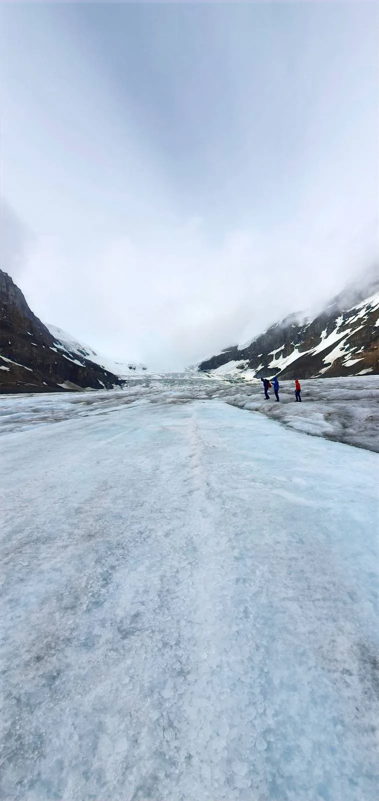A wide, icy glacier stretches between snow-covered mountains under a cloudy sky, with a few people walking in the distance.
