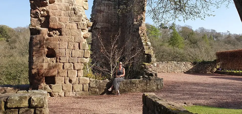 Woman in striped dress sits against ancient stone ruins with trees in the background. Sunny day in a natural, historic setting.
