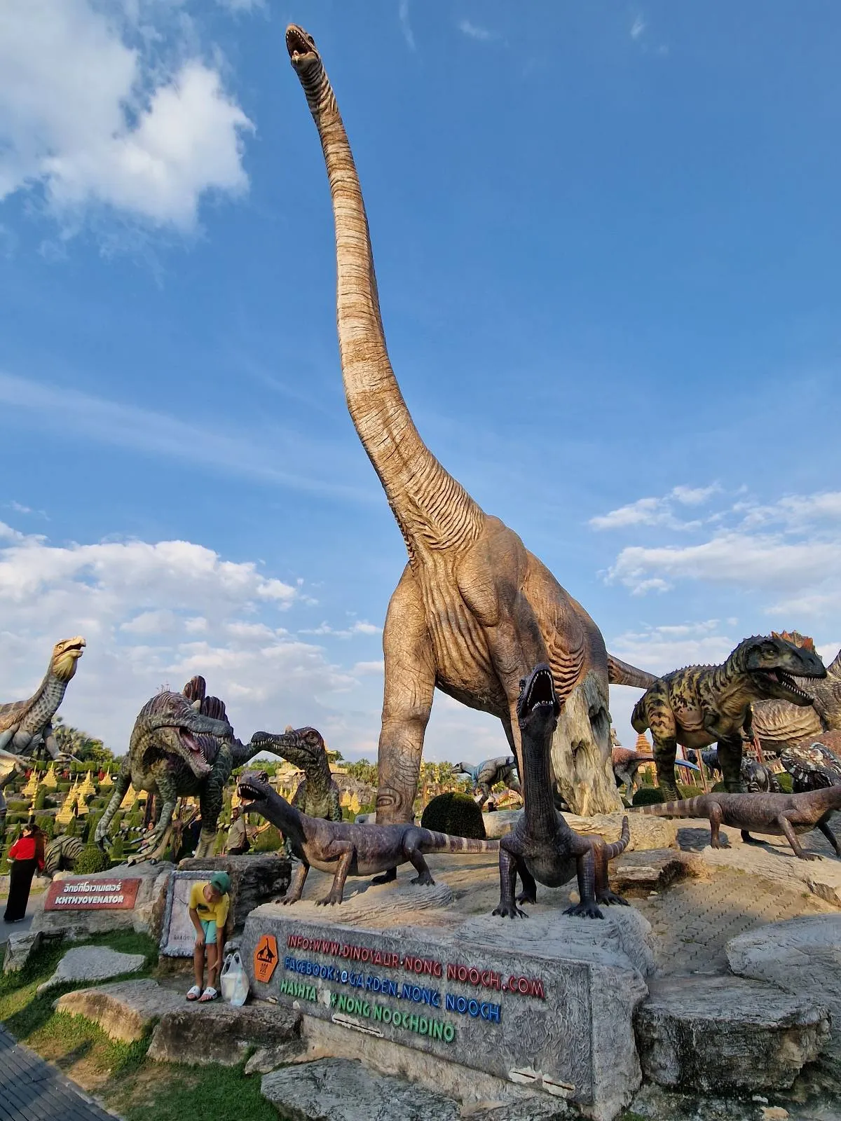 A large dinosaur statue with a long neck towers over other dinosaur sculptures in an outdoor park. A person stands nearby, providing a sense of scale. The sky is blue with scattered clouds. A stone sign is in the foreground.