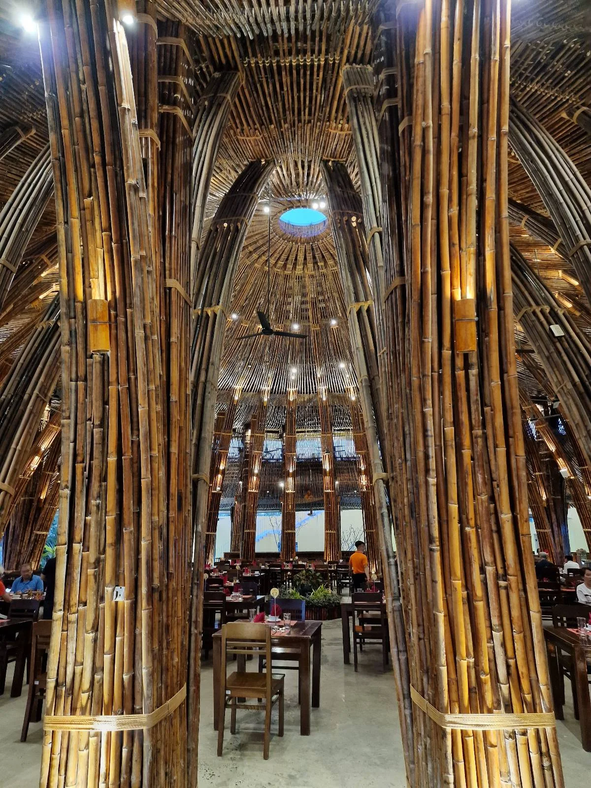 Interior of a restaurant with walls and ceiling constructed from bamboo poles, featuring dining tables and patrons.